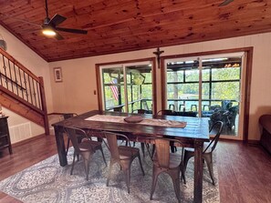 A dining area with a wooden table and chairs, a ceiling fan, and large windows with an American flag.
