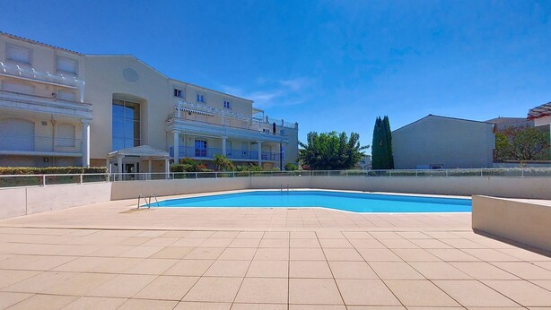 A swimming pool area with tiled flooring, a building with balconies, and a clear blue sky.