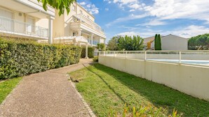 A pathway leading to a building with a white facade and balconies, surrounded by greenery and a clear sky.