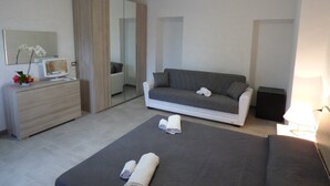Una camera da letto moderna con un divano grigio, un televisore su una cassettiera di legno, uno specchio e un letto con asciugamani arrotolati.