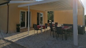 Una veranda coperta all'aperto con tavoli e sedie, una pianta in vaso e un edificio sullo sfondo.