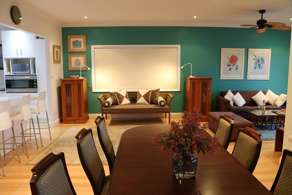 A dining area with a wooden table, chairs, and a vase of flowers. There is a sofa and a couch in the background. - Luxury Villa, Open Fire, Spa, Harbor Views, Pets, Babies, Surfing, Snorkeling (Ulladulla)
