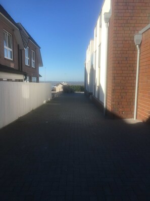 A narrow paved alleyway between buildings with brick and white siding, a white fence, and a clear blue sky.