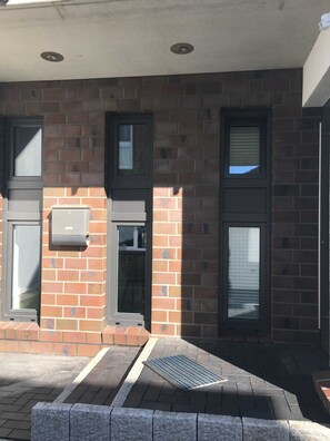 A building with a brick facade, two windows with blinds, a door, and a small air conditioning unit mounted on the wall. - Papen, A. Ferienwohnung An Bord - An Bord (Varel)