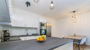 A modern kitchen with white cabinets, a stainless steel refrigerator, a built-in oven, and a fruit bowl on the counter. There is a dining table with chairs and pendant lighting above. - Holiday house SAN MICHELE (Vrh)
