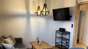 Un salon moderne avec une table basse en bois, un canapé recouvert d’un plaid, un téléviseur fixé au mur et un coin repas comprenant une table et des chaises.