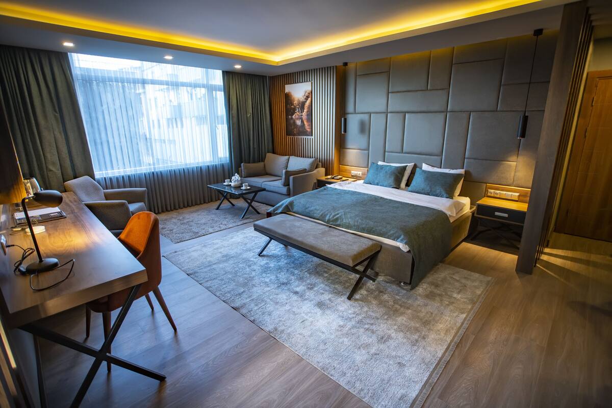 A modern hotel room with a large bed, a desk, a sofa, and a bench.