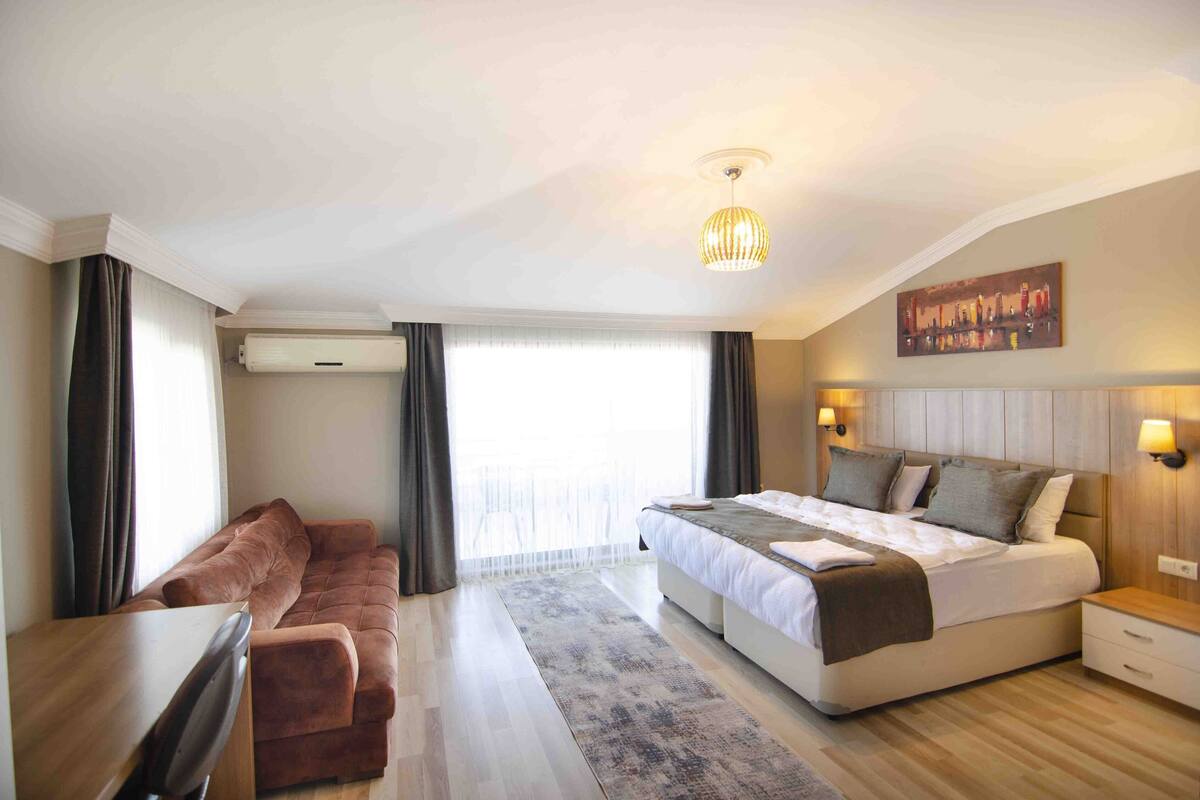 A modern hotel room with a large bed, a sofa, a desk, and a view of the city through the window.