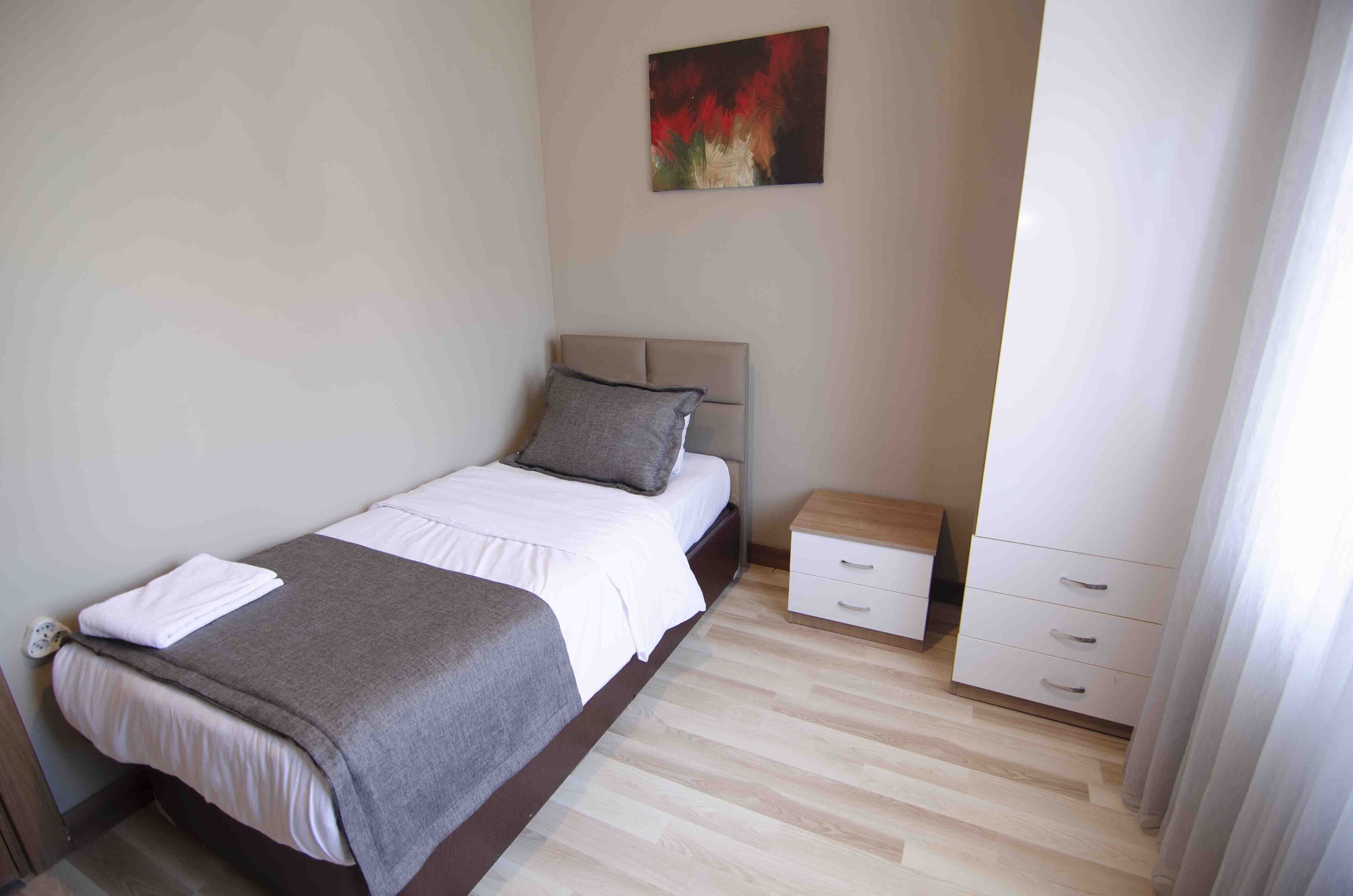 Una cama bien tendida con una manta gris y sábanas blancas, una mesita de noche con un cajón y un cuadro en la pared.