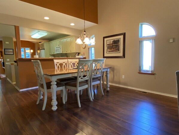 A dining area with a wooden table and chairs, a kitchen in the background, and a window with a view.