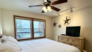 Un dormitorio con una cama grande, un ventilador de techo, un televisor sobre un aparador y una ventana con vistas a un balcón.