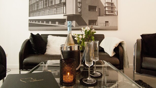 Una sala de estar moderna con una mesa de centro de vidrio, una botella de champán, una vela y dos copas de vino. En la pared, una fotografía en blanco y negro enmarcada de un edificio con la palabra "BAUHAUS".