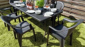 Mesa rectangular al aire libre con estructura metálica oscura, rodeada por seis sillas de plástico. La mesa está dispuesta con platos, tazones y vasos blancos, y presenta una jarra, una botella de vino y una copa de vino rosado.