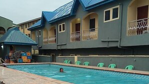 A swimming pool with a person swimming, surrounded by buildings with balconies and a covered area. - Perfecto Hotel Suites Lounge (Ibadan)