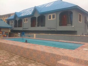 A building with a swimming pool, a tiled walkway, and a person in the pool. - Perfecto Hotel Suites Lounge (Ibadan)