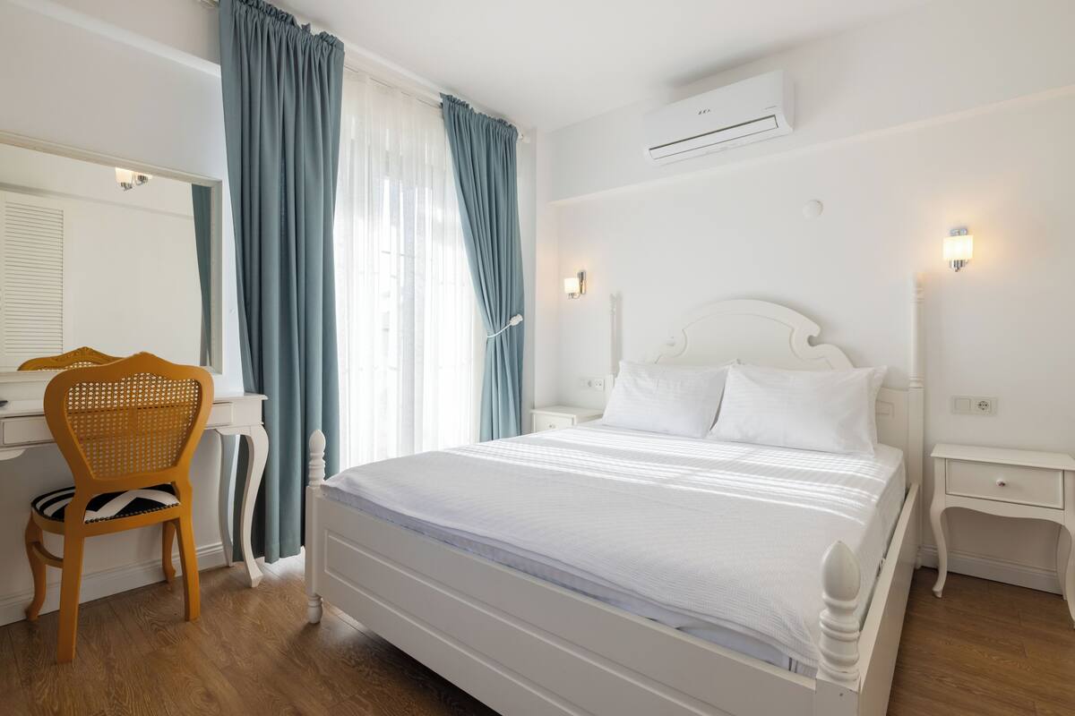 A hotel room with a white bed, a wooden chair, a desk, and a window with curtains.