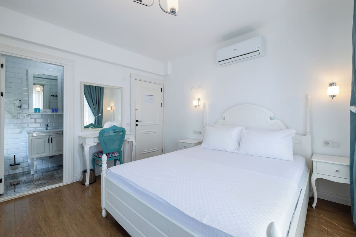 A white bedroom with a large bed, a desk with a chair, a mirror, and an air conditioning unit on the wall.