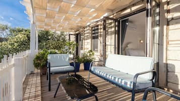 Een houten terras met een witte omheining, twee blauwe fauteuils met kussens, een bijzettafeltje en potplanten.