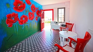 A room with a vibrant poppy mural, a tiled floor, a round table with chairs, and a window with a view. - Fitoz Otel (Bozcaada)