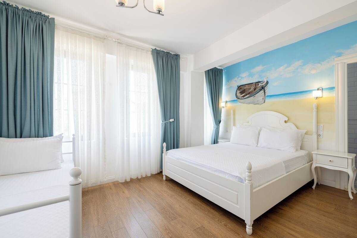 A bedroom with a bed, curtains, a nightstand, a wall clock, and a beach mural.