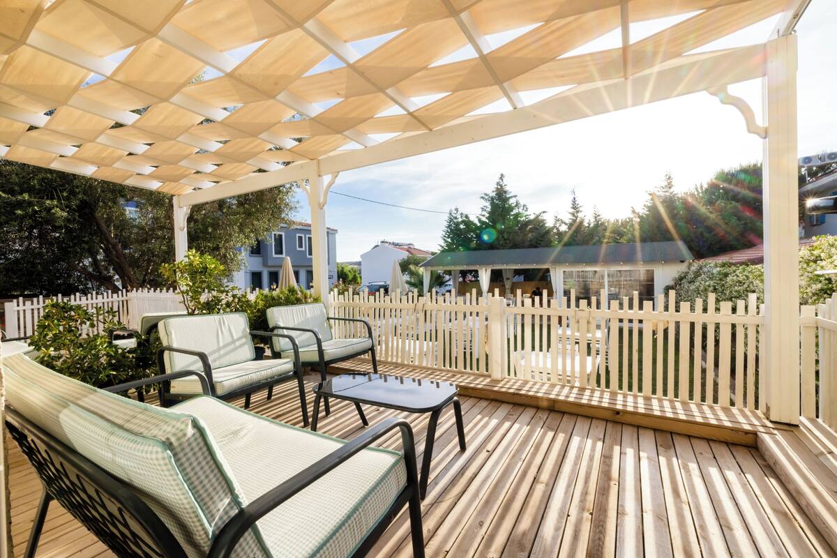 An outdoor patio with wooden flooring, a pergola, and a white picket fence.