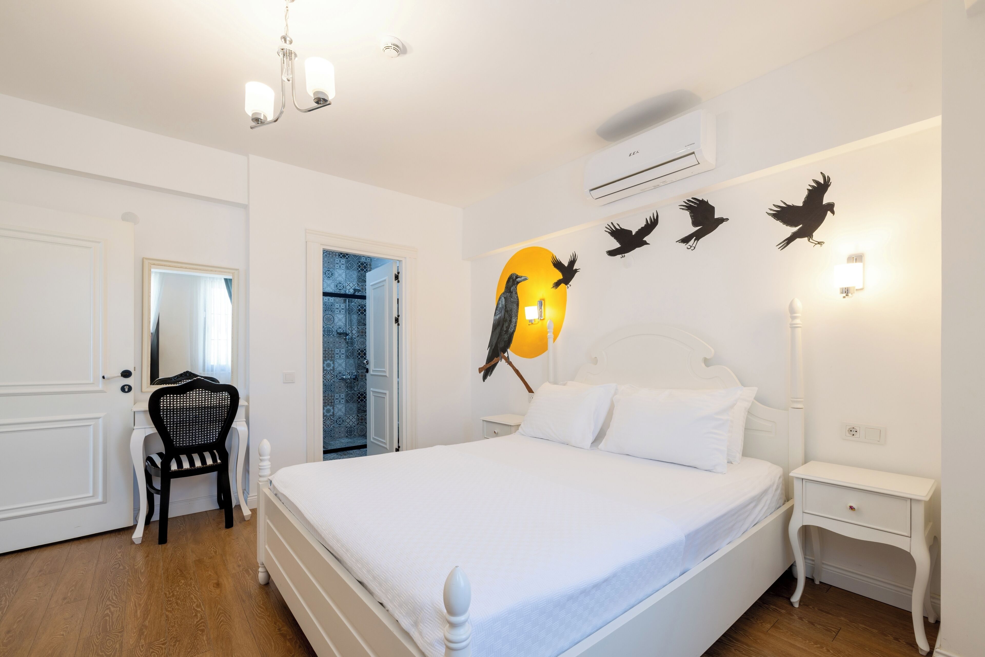 Quarto com cama branca, uma cadeira, mesa de cabeceira e arte na parede com pássaros.