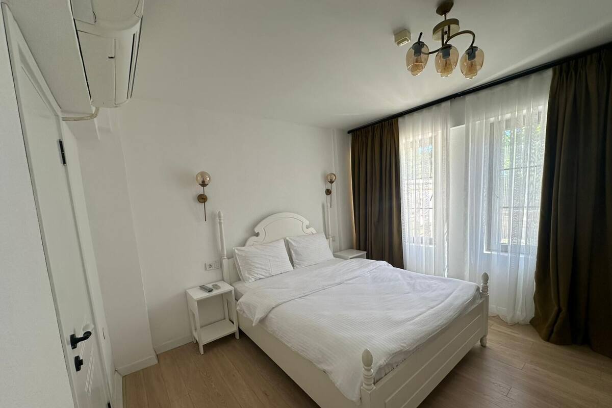 A bedroom with a bed, nightstand, wall sconces, a ceiling light fixture, and a window with curtains.