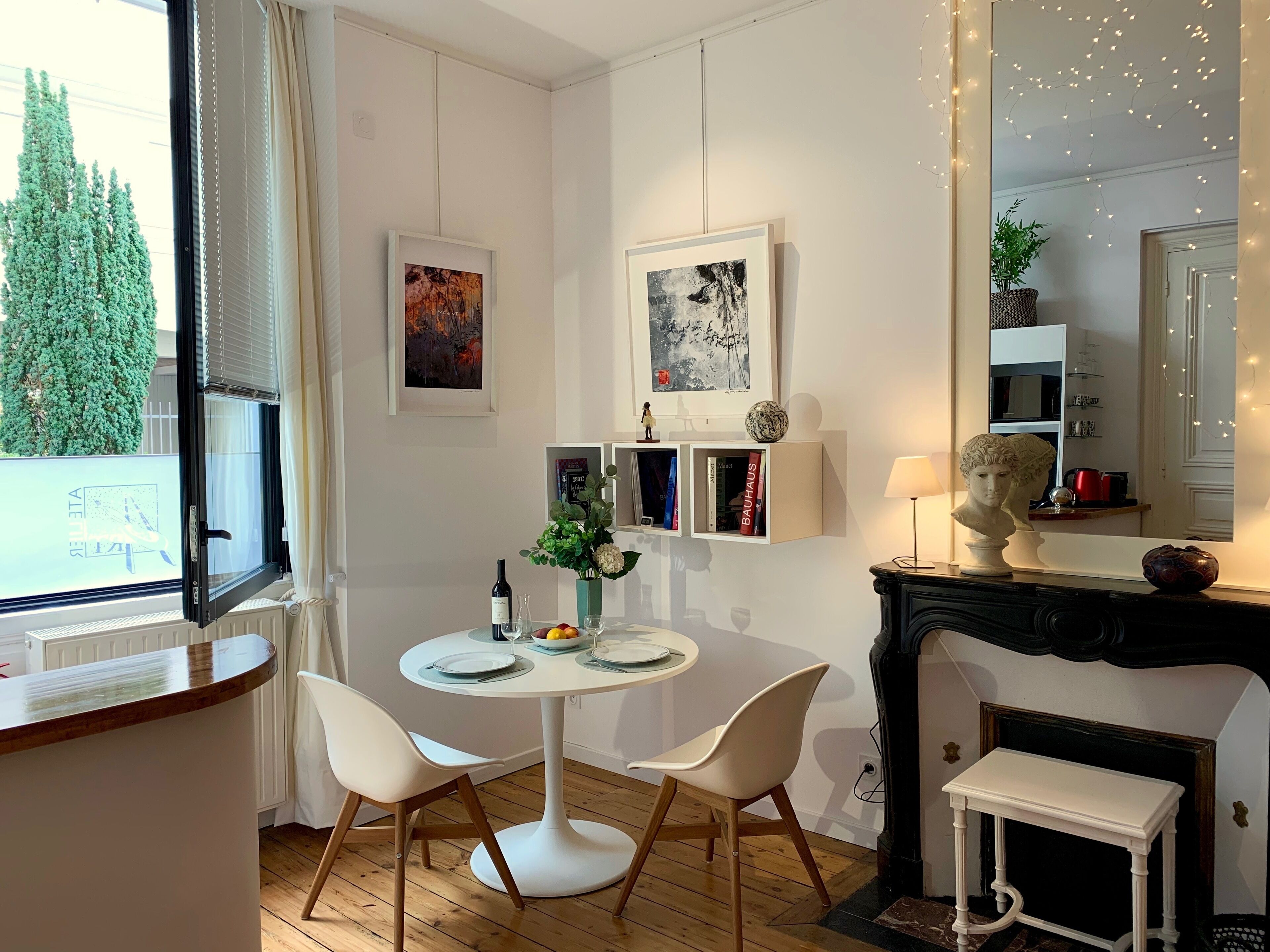 Un espace de salle à manger moderne avec une table ronde, des chaises blanches et une cheminée. Les murs sont ornés d’œuvres d’art encadrées, il y a une petite bibliothèque et une fenêtre avec des stores.