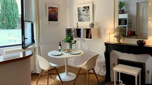 Un espace de salle à manger moderne avec une table ronde, des chaises blanches et une cheminée. Les murs sont ornés d’œuvres d’art encadrées, il y a une petite bibliothèque et une fenêtre avec des stores.