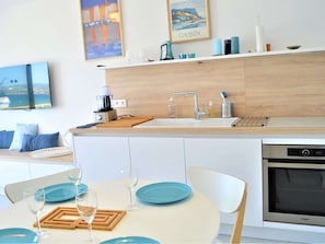 Uma cozinha moderna com forno embutido, cafeteira e pia. Há uma mesa de jantar com pratos azuis e copos de vinho.