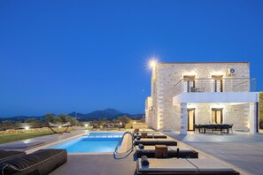 Un área junto a la piscina con sillones de descanso, una mesa de comedor y un edificio de piedra con balcón.