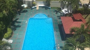 A rectangular swimming pool with a tiled deck, surrounded by palm trees and a red-roofed building.
