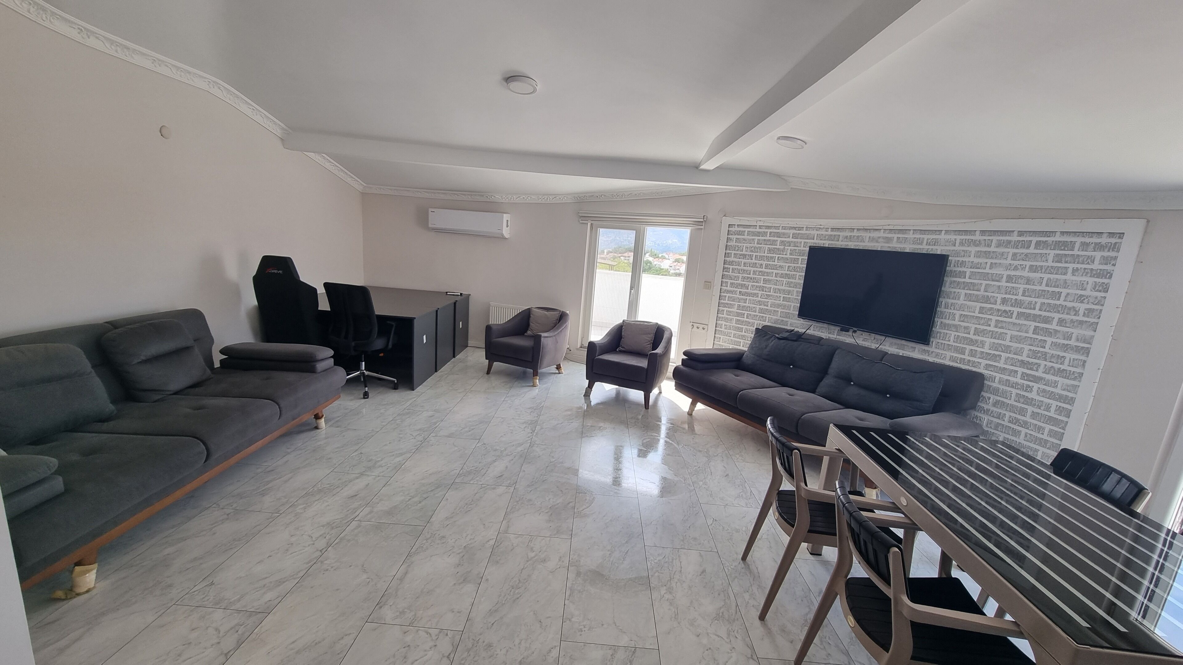 A spacious living area with a sofa, armchairs, a dining table, and a TV mounted on the wall.