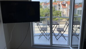 Una habitación con un ventanal grande, un televisor de pantalla plana en la pared y un balcón con mesa y sillas.