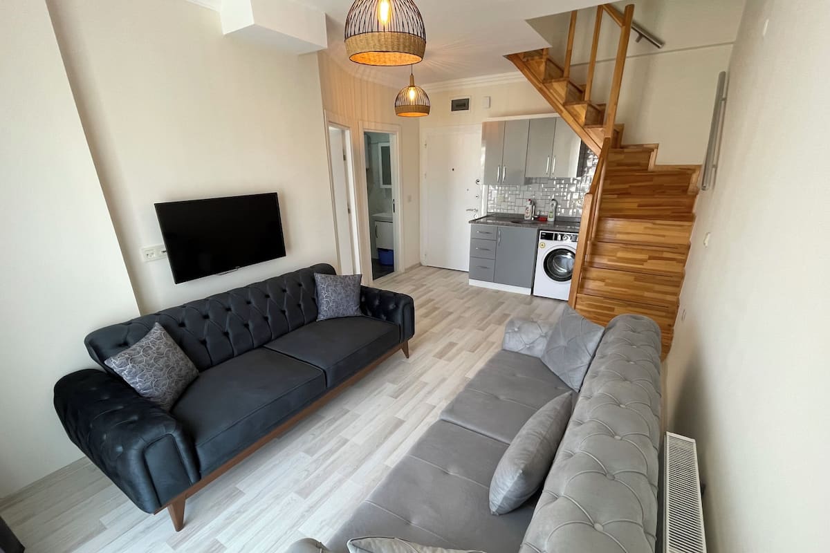 Ein modernes Wohnzimmer mit einer schwarzen Ledersofa und grauen Kissen, einem flachbildfernseher an der Wand und einer Treppe, die in einen Küchenbereich mit grauen Schränken und einer Waschmaschine führt.