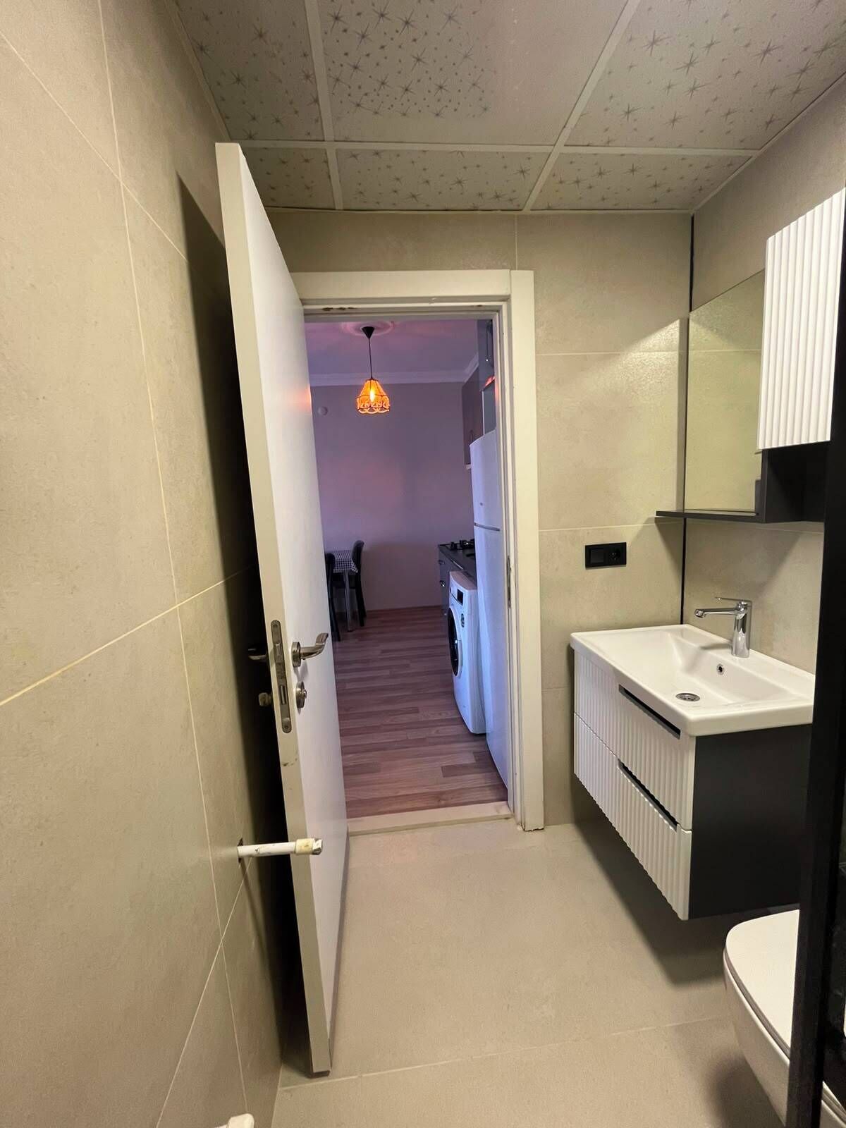 A narrow hallway leading to a kitchen with a washing machine, a sink, and a hanging light fixture.
