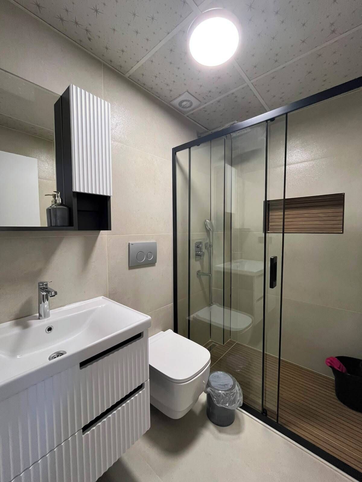 A modern bathroom with a white sink, a toilet, a glass shower enclosure, and a ceiling light.