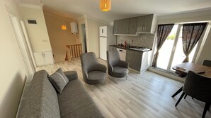 Una sala moderna con un sofá gris, dos sillones y una mesa de comedor con sillas. Se ve una cocina con armarios grises y una ventana al fondo.