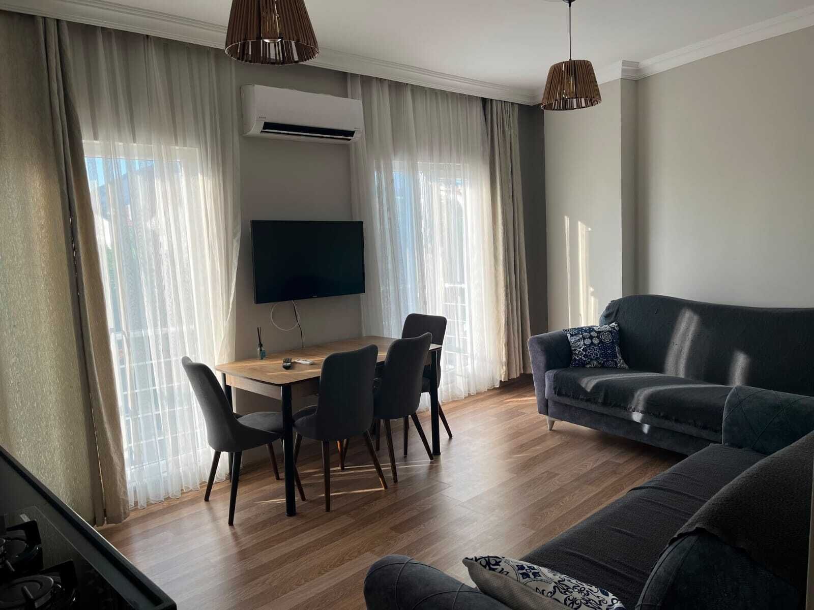 A modern living room with a flat-screen TV, a dining table with chairs, a sofa, and a staircase.