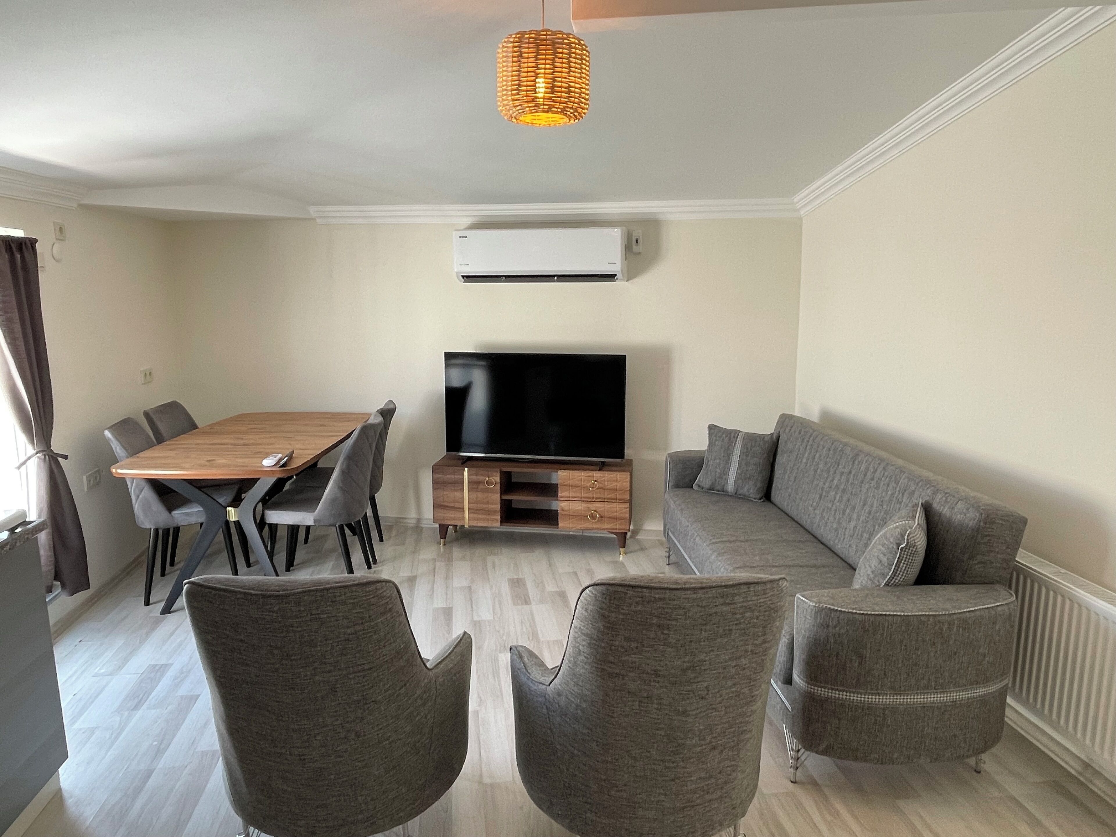 Un salon moderne avec une table à manger en bois, des chaises à revêtement gris, un canapé gris et un téléviseur à écran plat posé sur un meuble en bois.