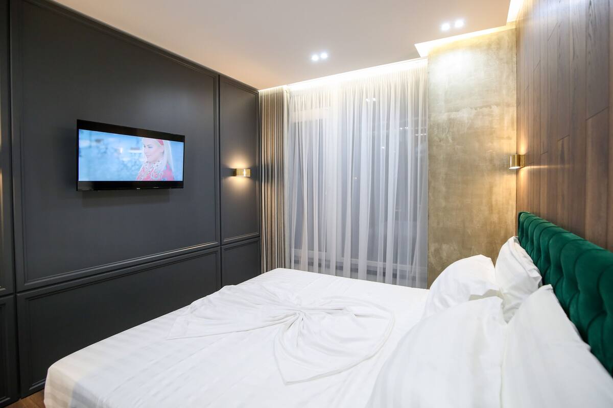A modern hotel room with a large bed, a flat-screen TV mounted on the wall, and sheer curtains.