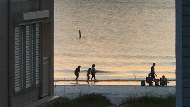 Solnedgang over kysten set fra en bygning med skodder, en strand i forgrunden, og et par personer der går en tur.