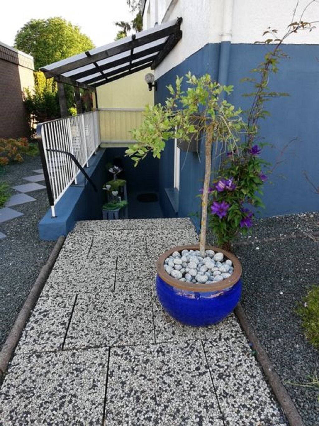 Ein kleiner Gartenbereich mit einem blauen Pflanzgefäß, das einen Baum und violette Blumen enthält, einem weißen Metallzaun und einem gepflasterten Weg, der zu einem Hauseingang führt.