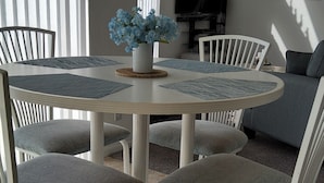 Une table de salle à manger ronde, entourée de chaises blanches, un vase de fleurs bleues et un téléviseur en arrière-plan.