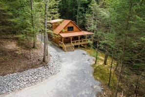 Une cabane avec une façade en bois, un porche et une allée de gravier, le tout entouré d’arbres.