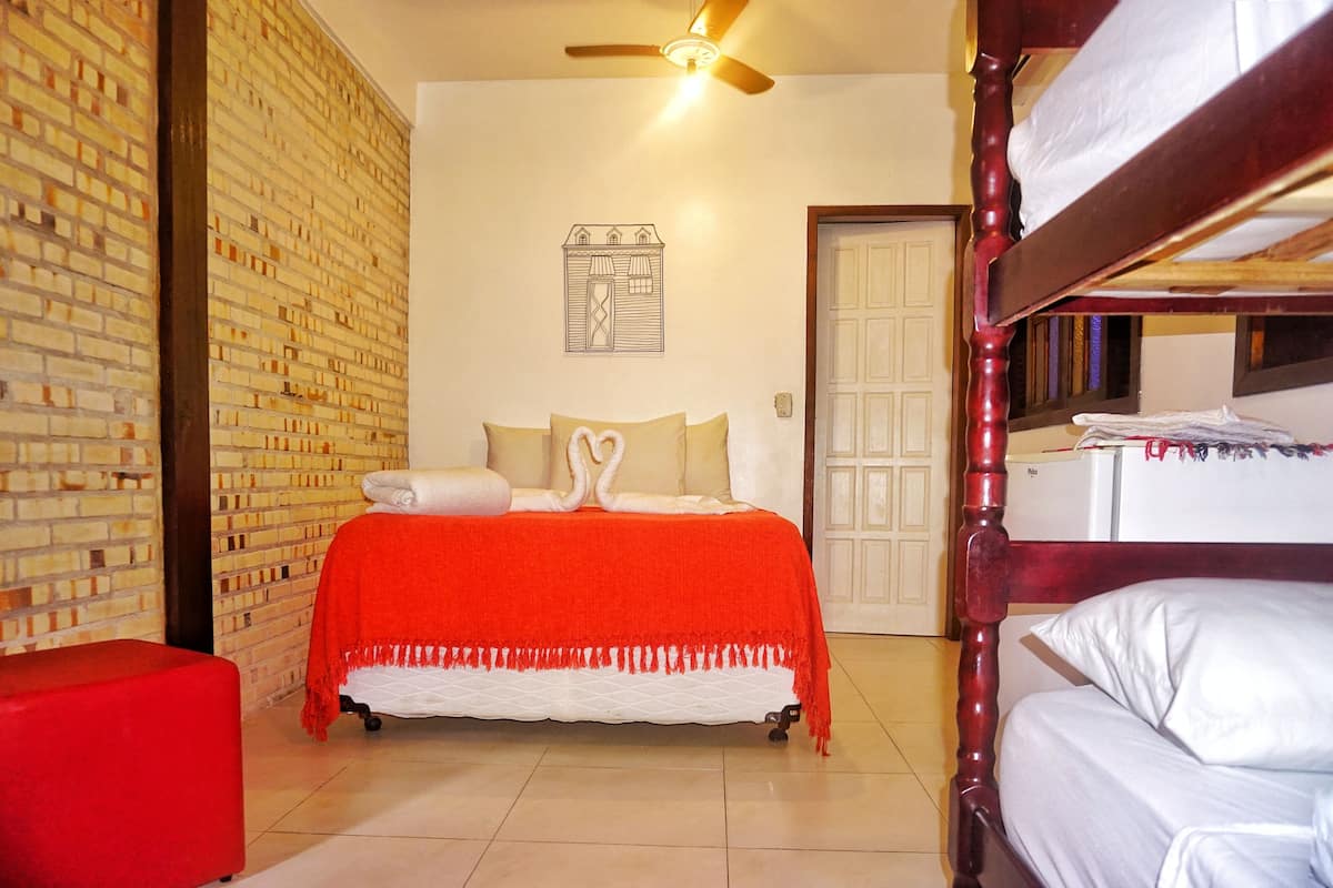 A bedroom with a bed, a red ottoman, a ceiling fan, and a brick wall.