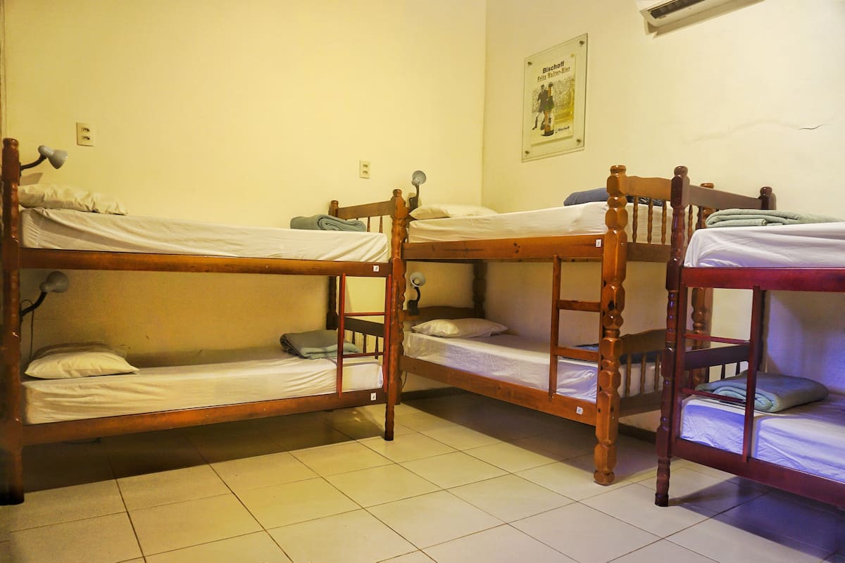 A dormitory room with three wooden bunk beds, a wall-mounted air conditioner, and a poster on the wall.