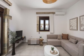 A living room with a sectional sofa, a flat-screen TV, a wooden sideboard, a small side table with a plant, and a window with curtains.