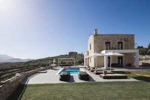 A pool area with lounge chairs, a pergola, and a stone building with a view of the mountains.