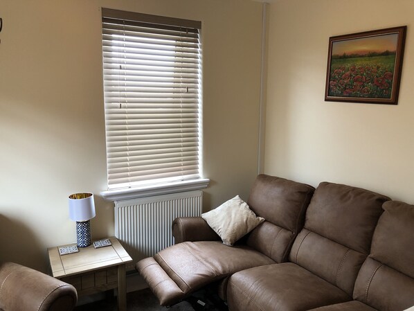 A living room with a brown leather sofa, a small table with a lamp, and a radiator. - Seacot Cottage in the heart of the Highlands (Clachnaharry)